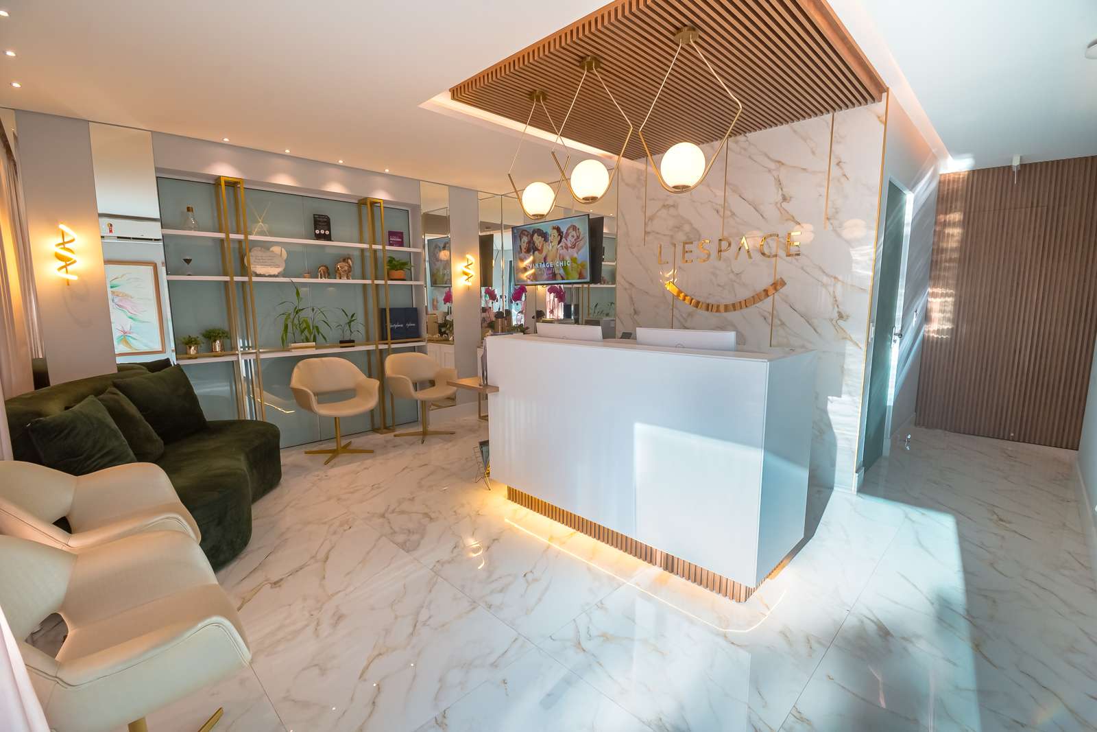 Recepção da L'Espace, com balcão branco iluminado, luminárias de globo e o logotipo dourado na parede de mármore